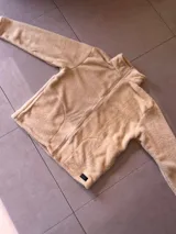 Buzo de poliéster color crema, con textura de sherpa, cuello alto y cierre frontal con cremallera.