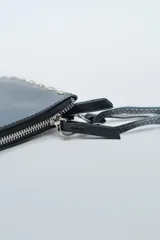 Cartera pequeña tipo clutch o bandolera de color negro, con borde decorado con tachas cuadradas plateadas. Posee cierre superior con cremallera y correa larga ajustable para llevar al hombro o cruzada.