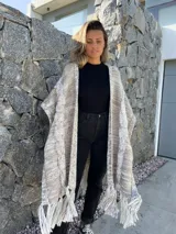 Ruana larga de lana merino color beige jaspeado, con diseño de trenzas y flecos en el borde inferior.