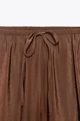Pantalón marrón de tejido fluido, tiro medio, cintura elástica ajustable con cordones, bolsillos laterales y falsos bolsillos de vivo en la espalda.
