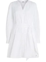 Vestido camisero corto color blanco, confeccionado en algodón orgánico. Presenta cuello en V, cierre frontal con botones, mangas largas abullonadas, cinturón de lazo ajustable en la cintura, bolsillos laterales y detalle de encaje calado en forma de escalera.