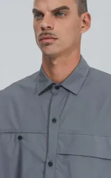 Camisa gris de manga corta confeccionada en poplin 100% algodón, con silueta oversize y dos bolsillos delanteros, uno con solapa y otro con ojal y botón.
