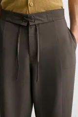 Pantalón marrón oscuro de corte amplio, con cintura ajustable con cordón, bolsillos plastrón delanteros y traseros, detalle de costuras frontales y cierre frontal con cremallera y gancho metálico oculto.