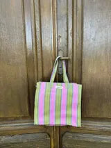 Bolso tipo tote bag con rayas verticales en tonos rosa, celeste y dorado. Tiene asas cortas rosas y etiqueta con la marca "AMÉN".