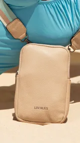 Phonebag color beige con textura granulada, logo "LINCOLN'S" grabado en el frente, cierre superior y bolsillo trasero tipo sobre. Incluye correa regulable y desmontable de galón con rayas en tonos beige, azul y verde.