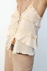Top de punto color crema con escote pico y tirantes finos. Detalle de volantes y cierre frontal con lazadas.