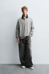Sudadera gris de corte holgado, con cuello polo negro, cierre frontal de tres botones y mangas largas con puños acanalados.
