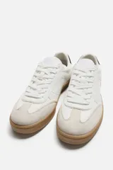 Zapatillas deportivas de estilo retro, color blanco con detalles en beige y negro. Capellada de cuero sintético con perforaciones laterales y puntera de gamuza. Suela de goma color caramelo.