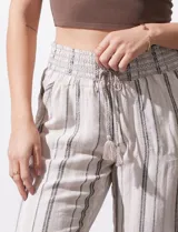 Pantalón de lino color beige con rayas verticales marrones, pretina ancha con punto smock y lazo ajustable, bolsillos verticales delanteros y bolsillos aplicados traseros.