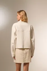 Chaqueta corta de jean color crudo, con cuello camisero, cierre frontal con botones metálicos y bolsillos de parche en el pecho con tachas metálicas.