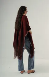 Ruana tejida a mano en lana merino 100%, de color marrón con detalles de costura en rojo en los bordes y terminación de flecos largos en el ruedo. Presenta una silueta amplia y caída natural, confeccionada artesanalmente por Manos del Uruguay.