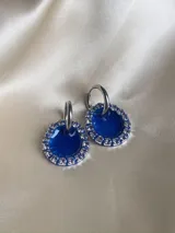 Aros colgantes con forma circular, hechos de resina color lila y borde de strass plateado. El aro es de acero quirúrgico plateado.