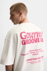 Camiseta blanca de corte relajado, con cuello redondo y mangas cortas. Presenta estampados combinados en contraste en la parte delantera y trasera con la frase "Gourmet Groove LLC".