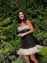 Vestido corto de lentejuelas negras con escote recto y tirantes finos. Presenta detalles de flecos de rafia en color beige tanto en el escote como en el ruedo.