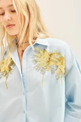 Camisa celeste de poplin con bordados de palmeras doradas en el frente, de corte amplio, con cuello y cartera.