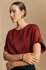 Top negro de manga corta con silueta amplia y caída fluida, confeccionado en un tejido liviano con leve brillo. Presenta un botón de nácar en la espalda con una pequeña abertura y mangas amplias tipo kimono.