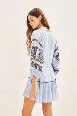 Vestido corto de bambula color celeste claro con bordados florales multicolor en el frente y mangas. Presenta escote redondo con cartera, cinturón de tela ajustable y volado en el ruedo.