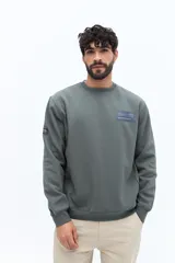 Buzo de algodón y poliéster color verde musgo, con cuello redondo y puños acanalados. Presenta un parche estampado rectangular con texto y un logo circular en el pecho.