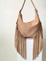 Bolso tipo hobo color tostado, hecho de cuero con flecos largos en el frente y dos bolsillos con cierre. Tiene una manija regulable con hebillas metálicas.
