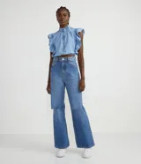 Camisa cropped de jeans celeste, sin mangas, con cuello alto, cierre frontal con botones, volantes en cuello y laterales, y elástico en la cintura.