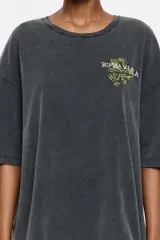 Camiseta de algodón orgánico color gris oscuro con efecto desgastado (acid wash). Presenta un corte oversize, escote redondo y un pequeño estampado gráfico en tono amarillo/verde en el pecho.