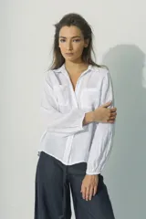 Camisa blanca de lino con cuello clásico, cierre frontal con botones, mangas largas con puños elásticos y dos bolsillos de parche en el pecho.
