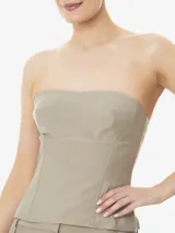 Top corset color beige, sin tirantes, confeccionado en tela ligera no elástica. Presenta escote recto, aberturas delanteras y traseras, largo corto, bajo asimétrico y cierre de cremallera en la espalda.