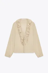 Camisa color beige de algodón con escote en V y volantes. Tiene mangas largas con botón en los puños y cierre con lazos en el frente.