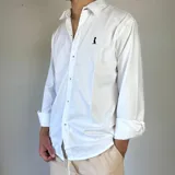Camisa blanca de algodón con cuello clásico, manga larga y cierre frontal con botones. Tiene un pequeño logo bordado en el pecho.