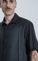 Camisa negra de manga corta con cuello clásico y cierre frontal con botones. Presenta una tira vertical cosida en el frente.