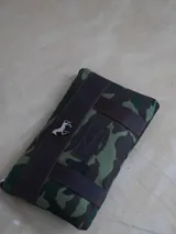 Sobre tipo cartuchera de lona con estampado camuflado en tonos verde y marrón, con terminaciones en cuero marrón. Tiene logo de la marca Sierra Mora y un caballo metálico.