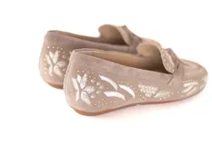 Mocasín de cuero gamuzado color avena, con bordados contrastantes, apliques metálicos y lazo decorativo en la capellada.