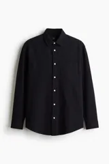 Camisa negra de sirsaca de algodón con cuello inglés, tapeta clásica, bolsillo superior de parche y canesú con pliegue de caja en la espalda. Mangas largas con tapeta de manga y botón, y cierre ajustable de botón en los puños. Bajo redondeado y corte holgado.