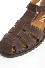 Sandalia cangrejera de gamuza color marrón oscuro, con puntera cerrada y tiras entrecruzadas. Posee tira de ajuste con hebilla metálica dorada en el tobillo y suela de goma plana.