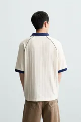 Polo color crema con cuello solapa azul marino, mangas cortas con ribete azul marino, bordado de texto en el pecho y bordado de logo en el abdomen.