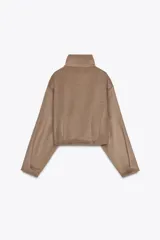 Cazadora corta color marrón claro, de cuello alto y manga larga. Tiene bolsillos laterales y cierre frontal con cremallera metálica.