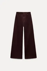 Pantalón de jean color marrón oscuro, de tiro alto y pierna ancha. Tiene bolsillos de parche en la parte trasera y cierre frontal con cremallera, botón y gancho metálico.