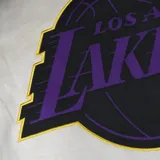 Campera bomber de algodón color crema con mangas de cuero sintético color violeta. Tiene el logo de Los Angeles Lakers bordado en el pecho y parches en los codos.