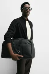 Bolso de viaje negro con compartimento principal con cierre de cremallera, bolsillo interior con cremallera, detalle de arnés en la parte frontal, dos asas de mano y asa de hombro ajustable y extraíble.