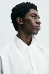 Camisa blanca de algodón popelín, corte relajado, con cuello solapa y pechera frontal plisada. Las mangas terminan en puños ajustables con lazos largos estampados en color marrón con patrón circular.