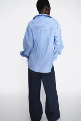Camisa celeste con rayas verticales blancas, de corte holgado, cuello solapa, escote en pico y mangas largas con puños abotonados.