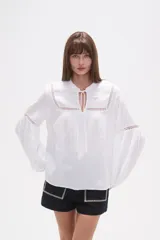 Blusa blanca de algodón con textura, manga larga abullonada y cuello mao con abertura frontal ajustable mediante cordones anudados. Presenta detalles de encaje calado en el canesú superior y en los puños.