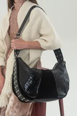 Cartera negra de cuero vacuno combinado: graneado, gamuza y grabado croco. Tiene silueta semicircular, correa ancha ajustable con herrajes metálicos y detalle de tachas. El interior está forrado en tela lisa y tiene un bolsillo con cierre.