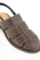 Sandalia tipo cangrejera de cuero marrón oscuro, con puntera cerrada tejida con tiras anchas y talón descubierto con tira ajustable y hebilla metálica. Posee suela de goma oscura y plantilla de cuero color camel con la marca grabada.