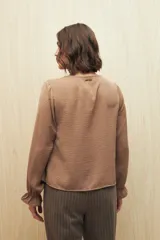 Camisa marrón de manga larga con escote en V y botones forrados al tono en el frente. Destaca por su textura sutil y por el detalle de frunces con volados en la parte inferior.