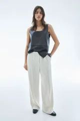 Pantalón de sarga color crudo con finas rayas verticales en gris, de tiro alto con cintura elástica y cierre decorativo. Tiene bolsillos laterales discretos y piernas anchas con pliegues cosidos adelante y pliegues planchados atrás.