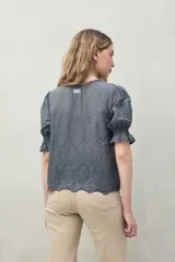 Blusa azul grisáceo de algodón con bordados frontales y terminaciones festoneadas. Tiene mangas cortas con frunces y cuello redondo con detalle de puntilla.