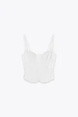 Top blanco estilo corsé con escote corazón, tirantes finos y bordados a tono. Presenta un bajo con volantes y cierre frontal con ganchos metálicos. La espalda es de tejido elástico tipo nido de abeja.