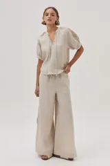 Blusa de lino color arena, de manga corta, con cuello redondo y lazo ajustable. Presenta bordados en las mangas y el escote.