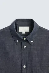 Camisa azul de algodón con textura chambray, corte regular fit, cuello abotonado, manga larga con puños abotonados, bolsillo de parche en el pecho y cierre frontal con botones.